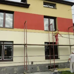 Dwóch mężczyzn na rusztowaniu podczas prac przy elewacji dwukondygnacyjnego domu z oknami o brązowych ramach, gdzie fragment ściany jest pomalowany na czerwono, a pozostała część na kremowo-żółty...