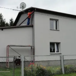 Dwupiętrowy dom po ociepleniu, z pracownikiem w pomarańczowej koszulce na dachu, poprawiającym rynnę, widok zza metalowego płotu z kwiatami.