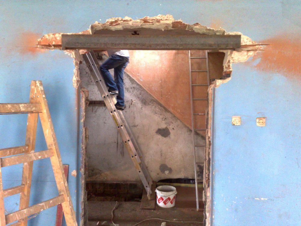 Pracownik na drabinie, montaż stalowej belki nad otworem w ścianie podczas prac remontowych wewnątrz budynku. Widoczne dwie drabiny, fragment niebieskiej ściany i uszkodzony mur.