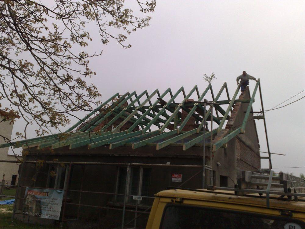 Budynek w trakcie rozbudowy: widoczna konstrukcja dachu z zielonych belek, murarz na szczycie ściany, fragment żółtego samochodu na pierwszym planie, pochmurne niebo w tle.