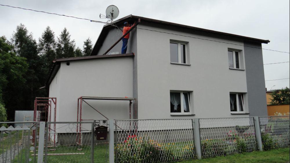 Dwupiętrowy dom po ociepleniu, z pracownikiem w pomarańczowej koszulce na dachu, poprawiającym rynnę, widok zza metalowego płotu z kwiatami.