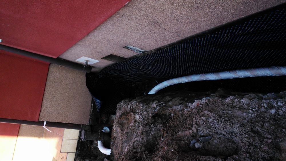 Fundament budynku zabezpieczony czarną folią izolacyjną, widoczny drenaż i fragment ocieplenia oraz rury.