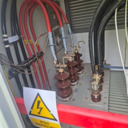 Intek-Tech Technika Instalacji Prażuch Piotr - Wnętrze szafy elektrycznej z grubymi kablami w kolorach czarnym i czerwonym, brązowymi izolatorami i żółtym znakiem ostrzegawczym 'Pod Napięciem'.