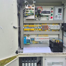 Intek-Tech Technika Instalacji Prażuch Piotr - Otwarta szafa sterownicza z widocznymi elementami instalacji elektrycznej: bezpieczniki, przewody, sterownik, akumulatory. Układ schludny, zorganizowany.