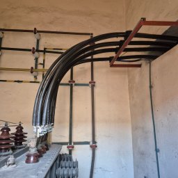Intek-Tech Technika Instalacji Prażuch Piotr - Wnętrze stacji transformatorowej: grube kable w czarnej izolacji, ceramiczne izolatory, metalowa konstrukcja wsporcza, ściany z surowego betonu. Widok z dołu.