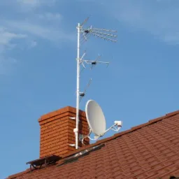 Antena satelitarna i antena Yagi-Uda zamontowane na kominie z czerwonej cegły na tle błękitnego nieba.