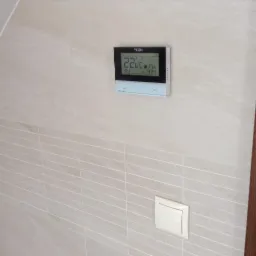 Elegancki, beżowy termostat TECH na ścianie z płytek, wyświetlający temperaturę 22.6°C i godzinę 14:24. Obok włącznik światła.