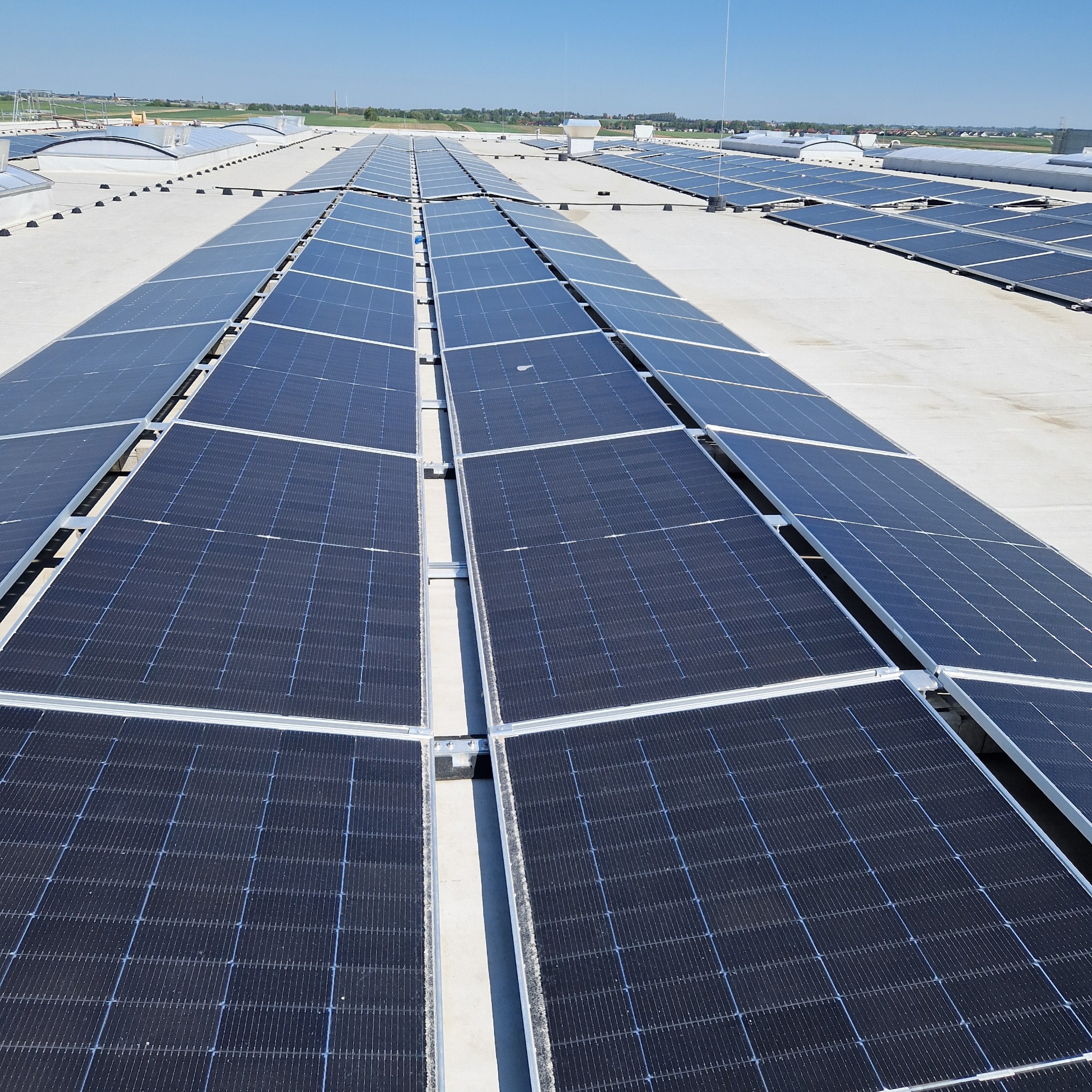 Rozległa instalacja paneli słonecznych na dachu budynku w słoneczny dzień. Widoczne rzędy paneli i elementy konstrukcyjne. Tło stanowi błękitne niebo i odległy krajobraz.