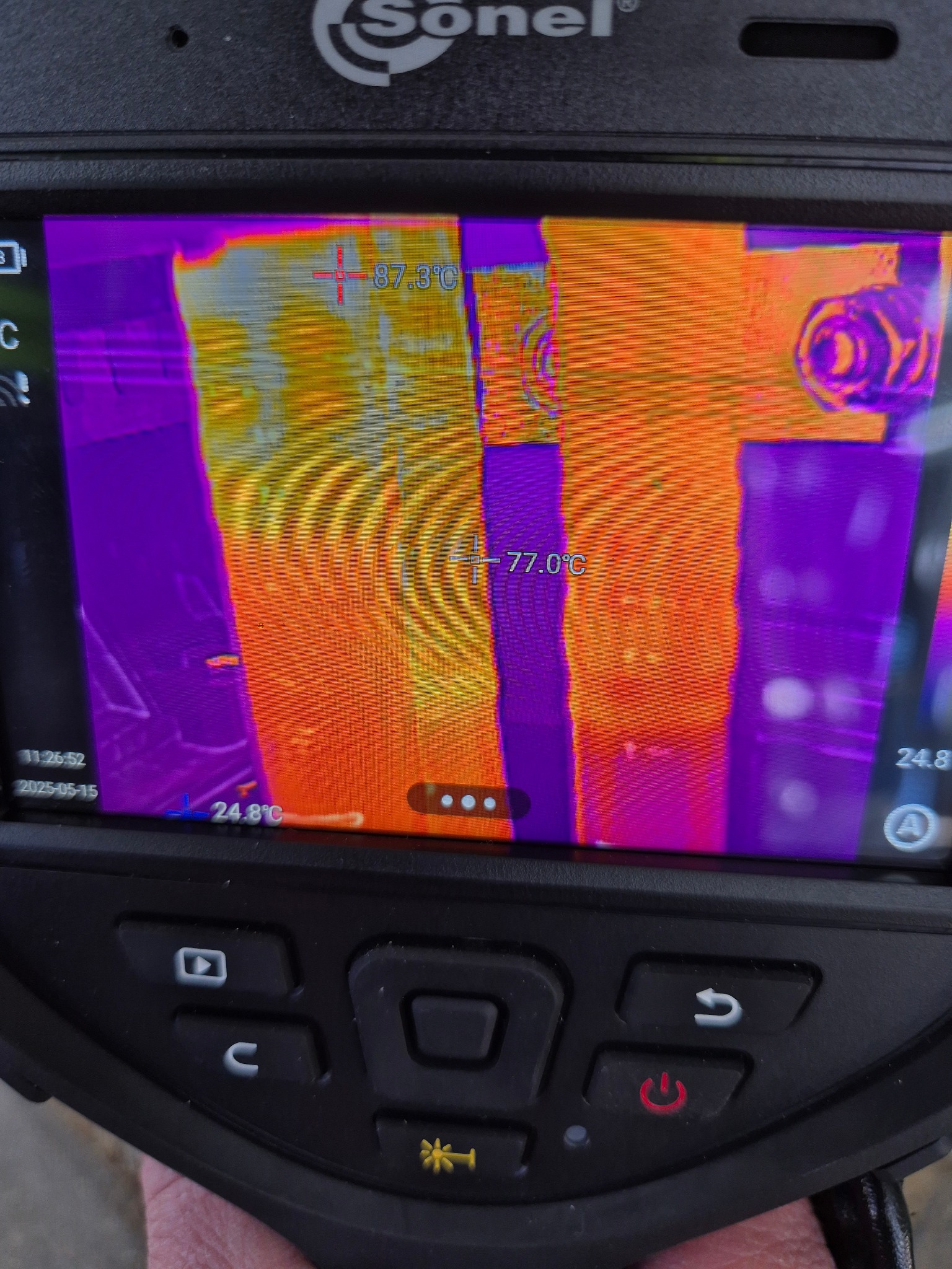 Ekran kamery termowizyjnej Sonel pokazuje rozkład temperatur na elementach instalacji, z zaznaczonymi punktami pomiarowymi 87.3°C i 77.0°C. Widoczne przyciski urządzenia.