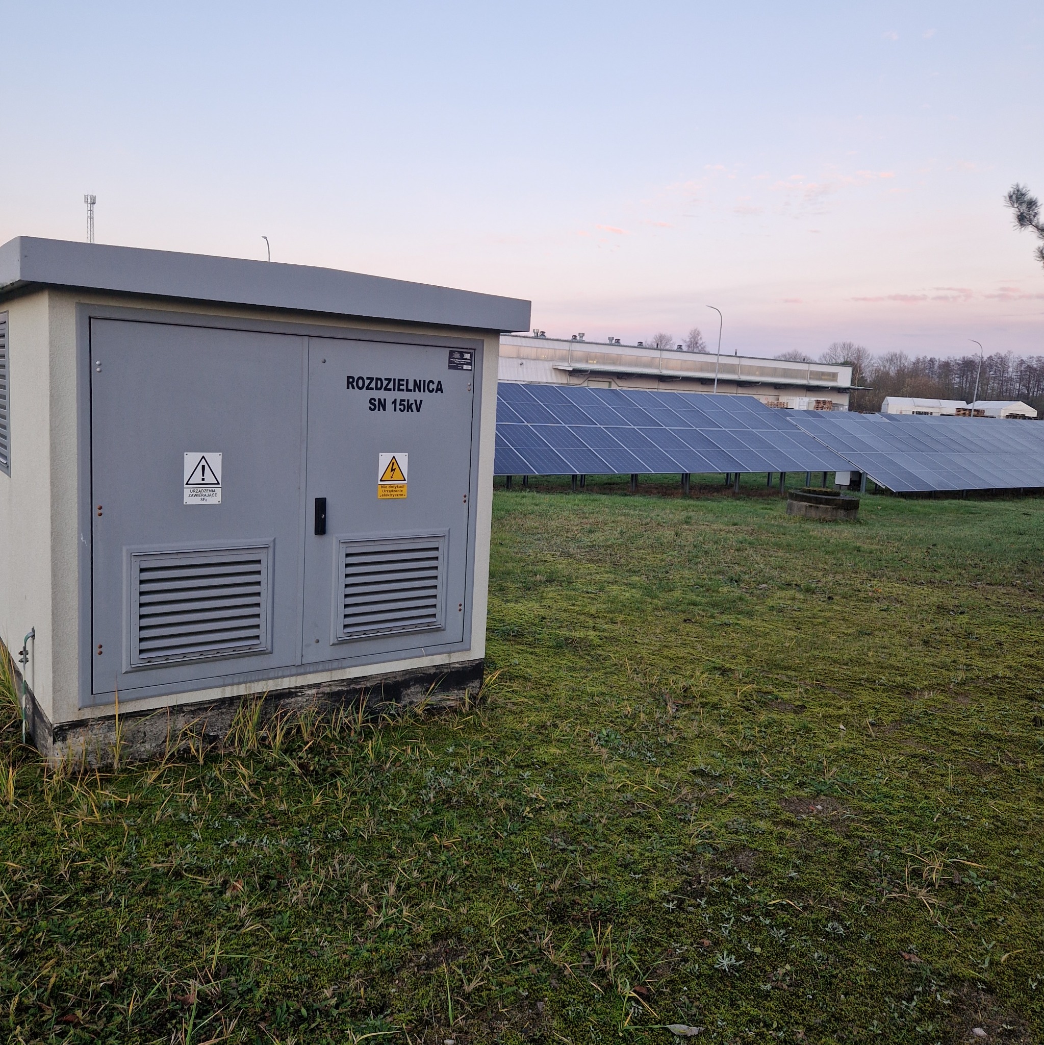 Szara rozdzielnica SN 15kV na tle farmy paneli słonecznych o zmierzchu. Tabliczki ostrzegawcze na drzwiach. Widok na zielony teren i budynki w oddali.