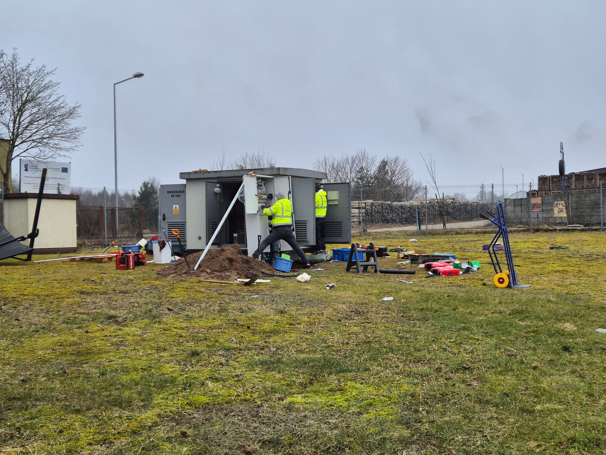 Pracownicy w żółtych kamizelkach serwisują rozdzielnicę elektryczną na zewnątrz, narzędzia i sprzęt rozłożone na trawie, pochmurne niebo w tle.