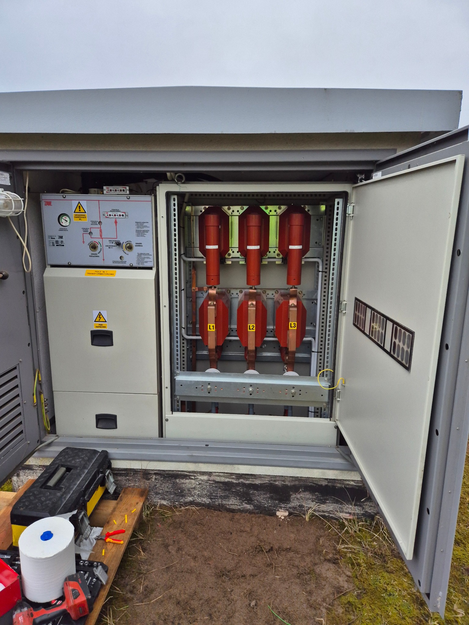 Otwarta szafa elektryczna z widocznymi zabezpieczeniami i elementami instalacji. Na zewnątrz skrzynka narzędziowa i akcesoria.