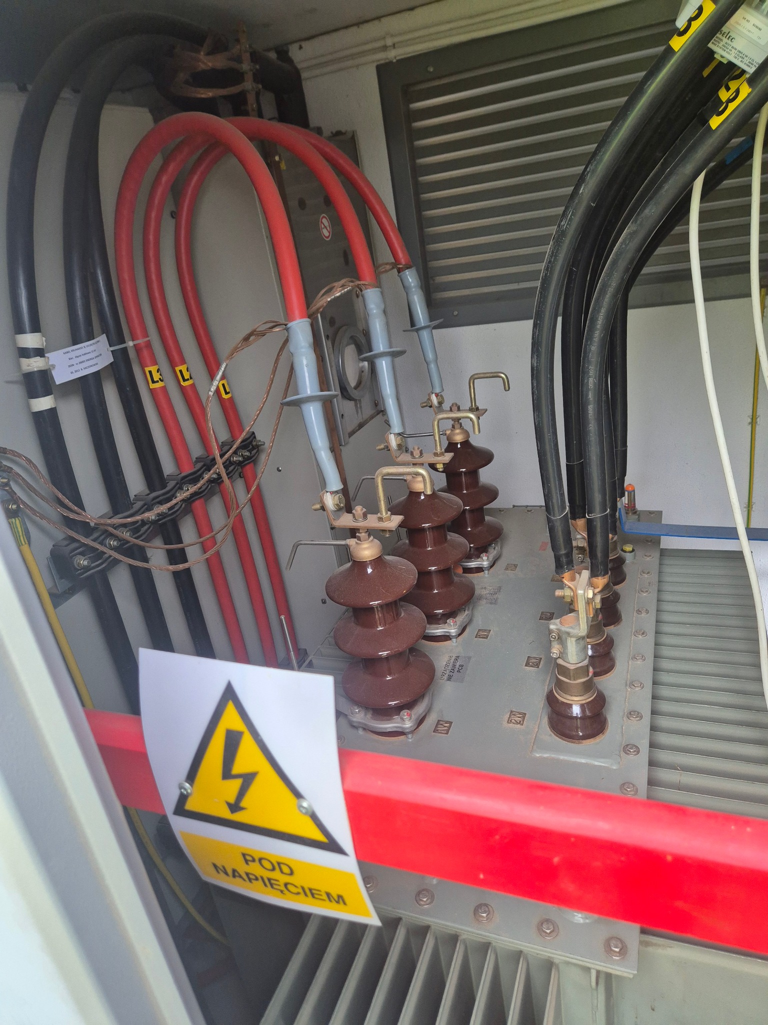 Wnętrze szafy elektrycznej z grubymi kablami w kolorach czarnym i czerwonym, brązowymi izolatorami i żółtym znakiem ostrzegawczym 'Pod Napięciem'.