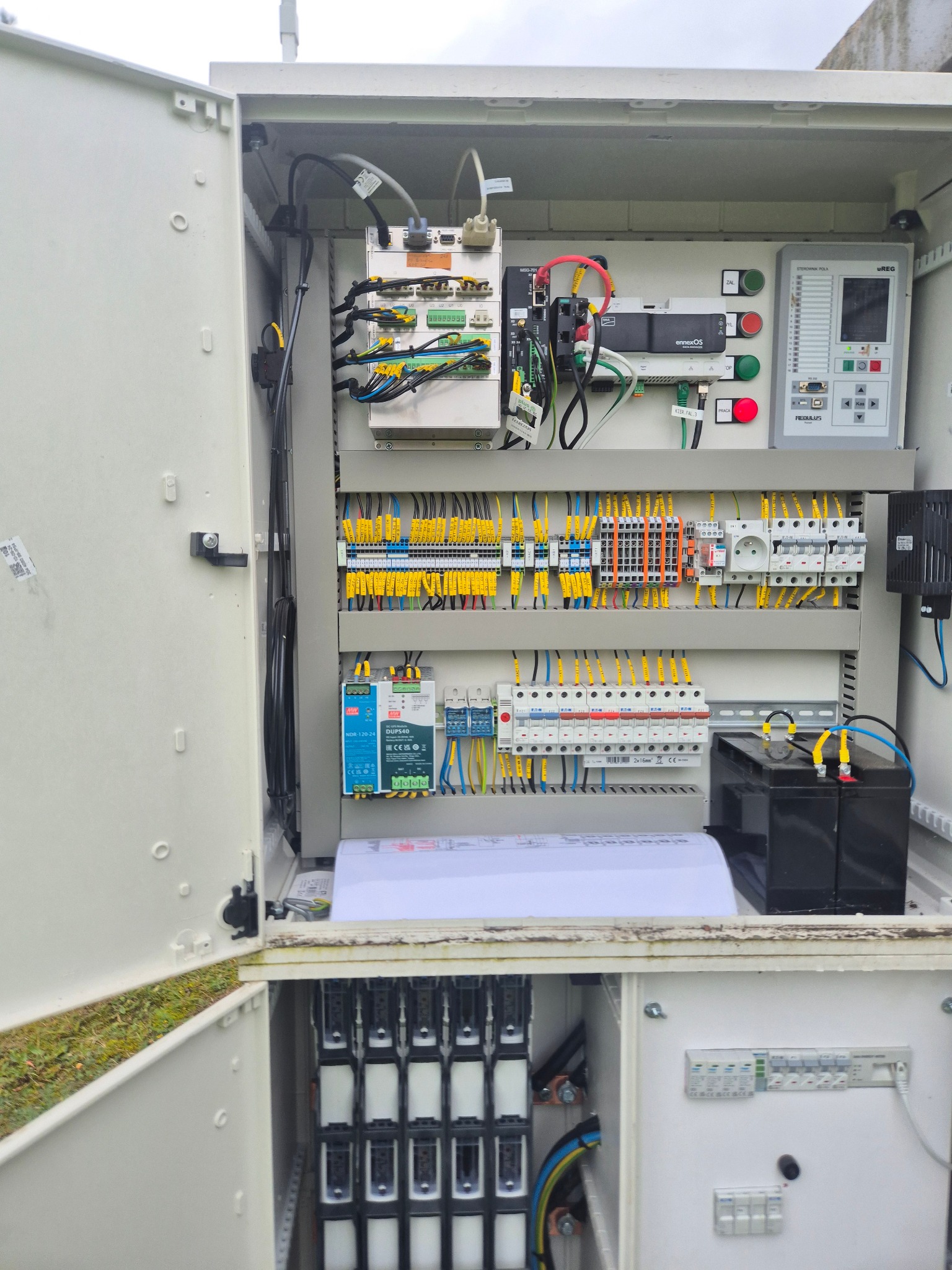 Otwarta szafa sterownicza z widocznymi elementami instalacji elektrycznej: bezpieczniki, przewody, sterownik, akumulatory. Układ schludny, zorganizowany.