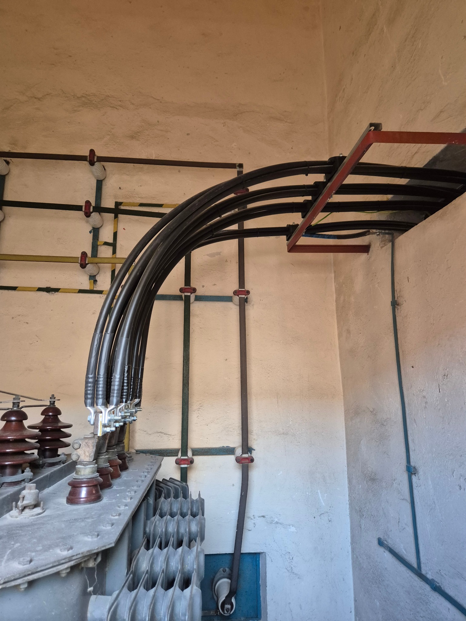 Wnętrze stacji transformatorowej: grube kable w czarnej izolacji, ceramiczne izolatory, metalowa konstrukcja wsporcza, ściany z surowego betonu. Widok z dołu.