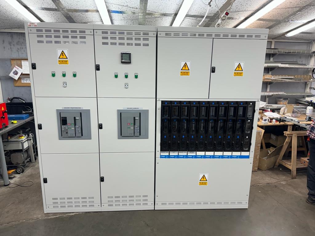 Rozdzielnica elektryczna w obudowie, z widocznymi zabezpieczeniami, wyłącznikami i oznaczeniami ostrzegawczymi. Widoczne elementy sterujące i pomiarowe.