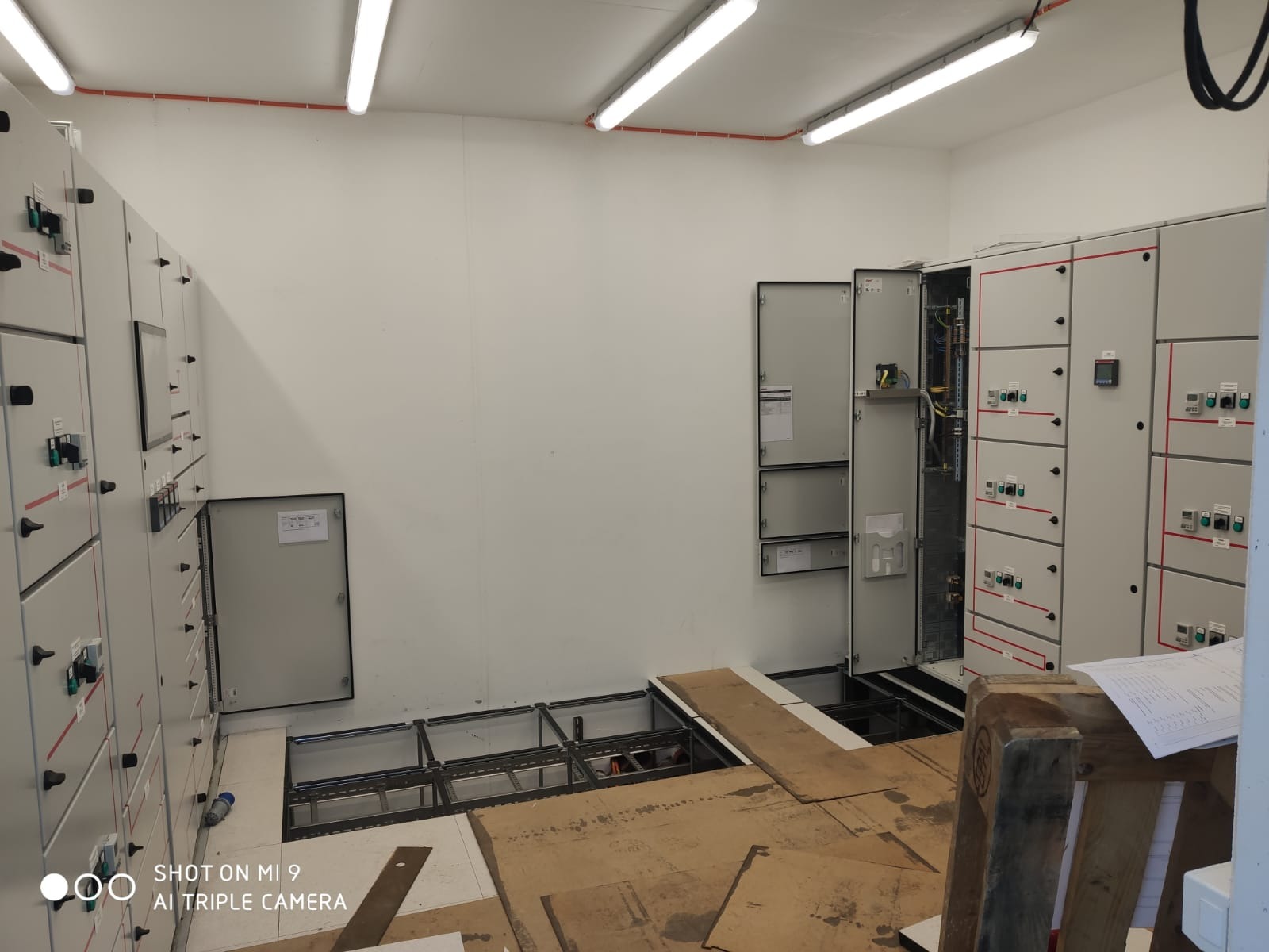 Wnętrze pomieszczenia z rozbudowaną instalacją elektryczną: szafy sterownicze z otwartymi drzwiami, kable i przewody, podniesiona podłoga z widoczną konstrukcją nośną.