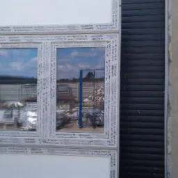 Trzy białe ramy okienne z szybami odblaskowymi, ułożone jedna na drugiej, widoczne logo Salamander i System Brügmann, tło z elementami budowlanymi i szarą ścianą.