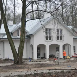 Nowoczesny, biały dom z szarym dachem z blachy, widoczny pracownik w pomarańczowej kamizelce, otoczenie w stanie surowym, drzewa bez liści