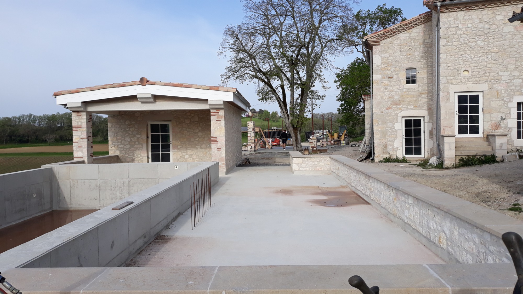 Budowa domu z kamienia: widok na budynek główny i aneks z basenem w trakcie prac wykończeniowych na zewnątrz, widoczne zbrojenia, teren budowy i maszyny w tle.