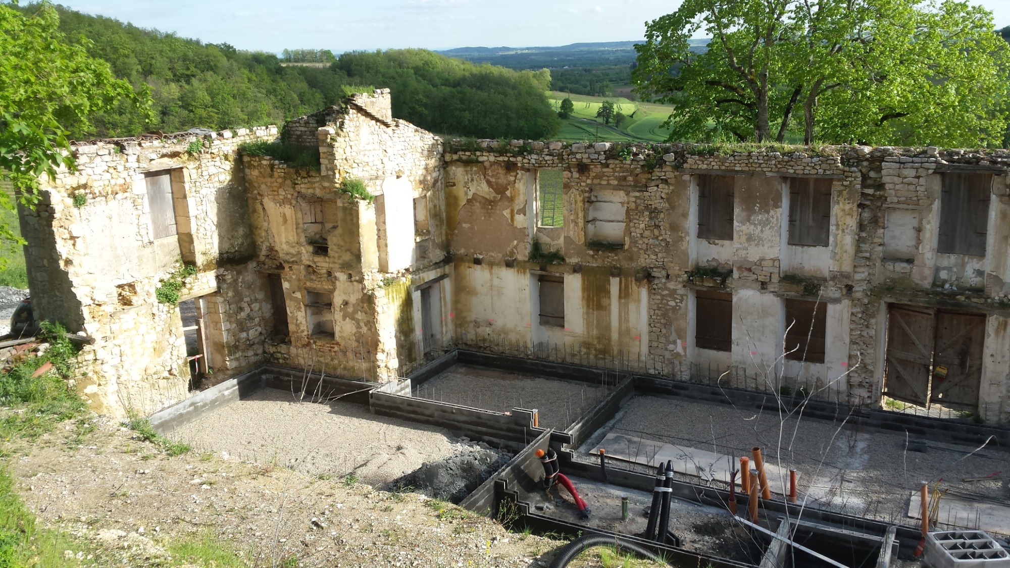 Ruiny kamiennego budynku z odsłoniętymi fundamentami i nową konstrukcją szalunkową, widok na zielone wzgórza w tle.