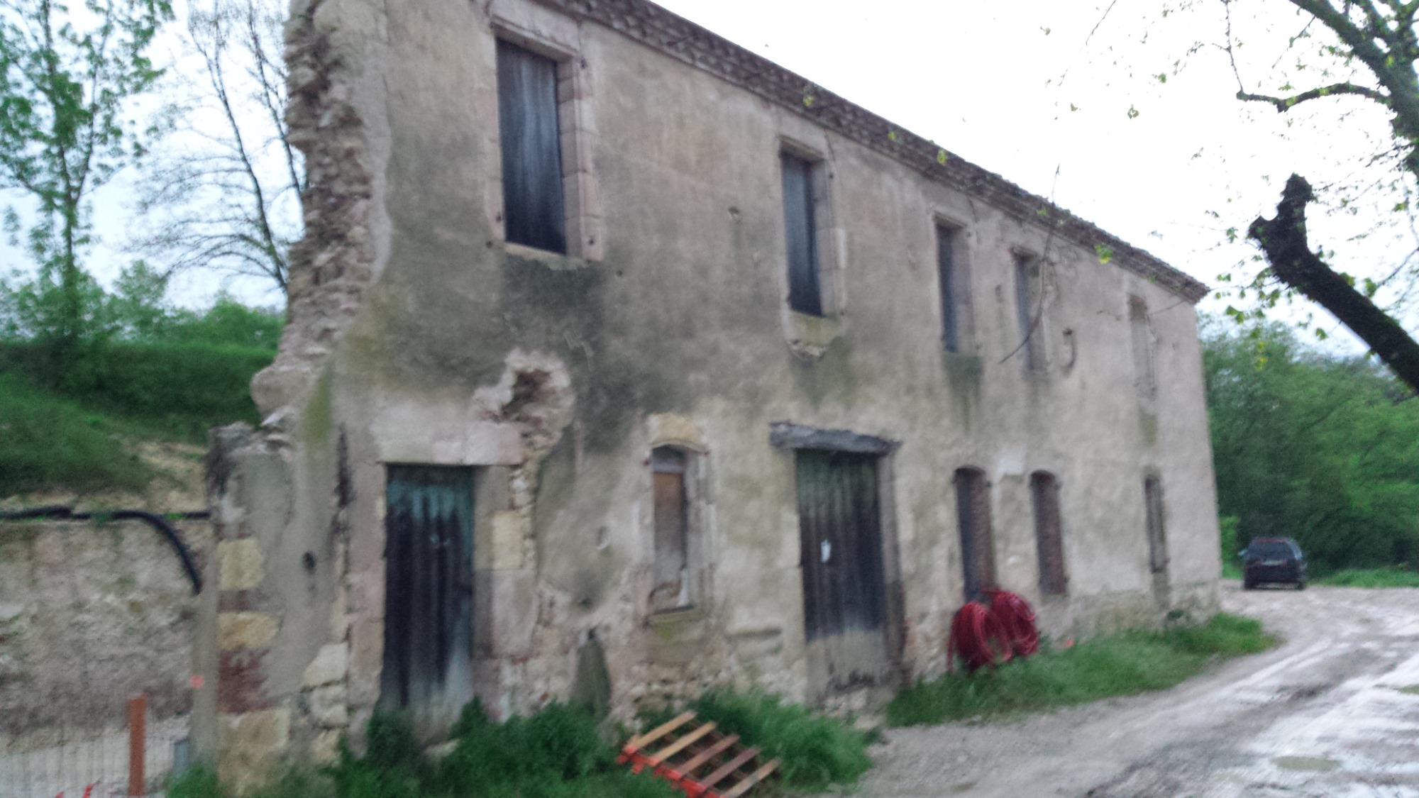 Zrujnowany, dwukondygnacyjny budynek z częściowo zawalonym narożnikiem, ukazujący odsłoniętą konstrukcję murowaną, z drewnianymi, zamkniętymi okiennicami i drzwiami, otoczony zielenią i błotnistą...