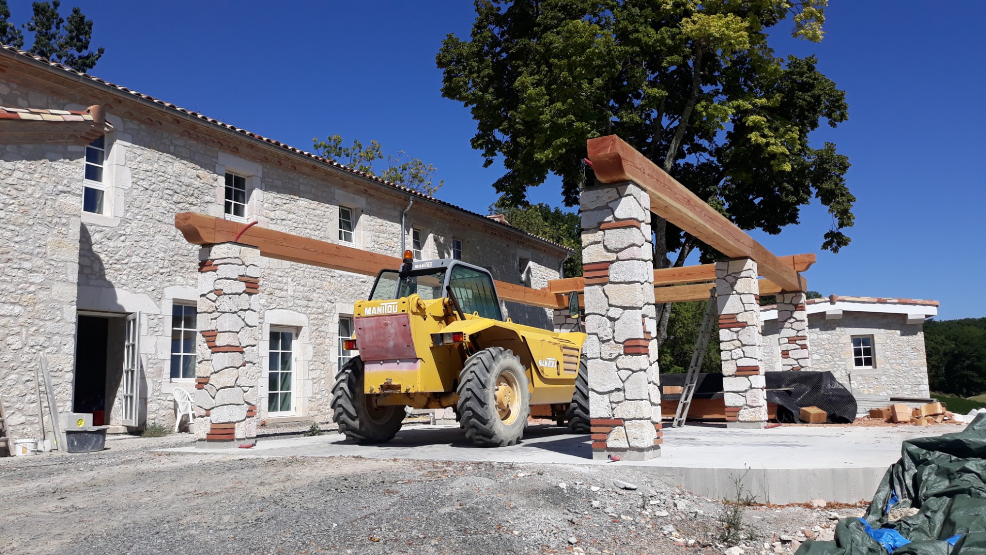 Wznoszenie kamiennego domu z elementami drewnianymi, widoczny podnośnik Manitou i rusztowanie, etap budowy w słoneczny dzień.