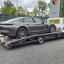 PHU Hubert Banat - Szary Porsche na lawecie pomocy drogowej, na tle salonu Porsche Centrum Łódź. Widoczny numer telefonu do pomocy drogowej i zaparkowany czarny samochód.