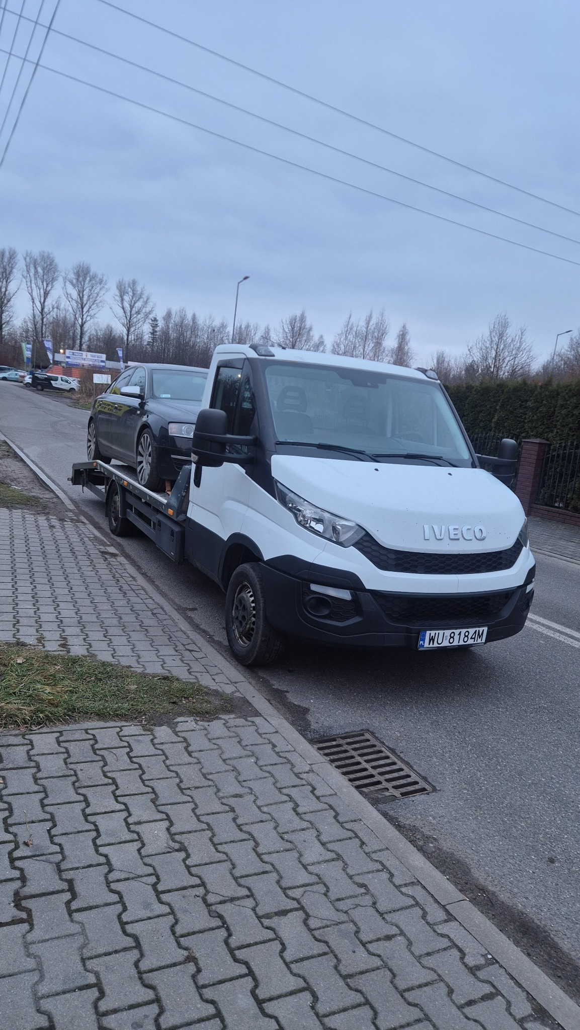 Biała laweta Iveco z czarnym samochodem osobowym na platformie, zaparkowana na poboczu drogi w pochmurny dzień. Widoczny fragment chodnika i drzewa w tle.