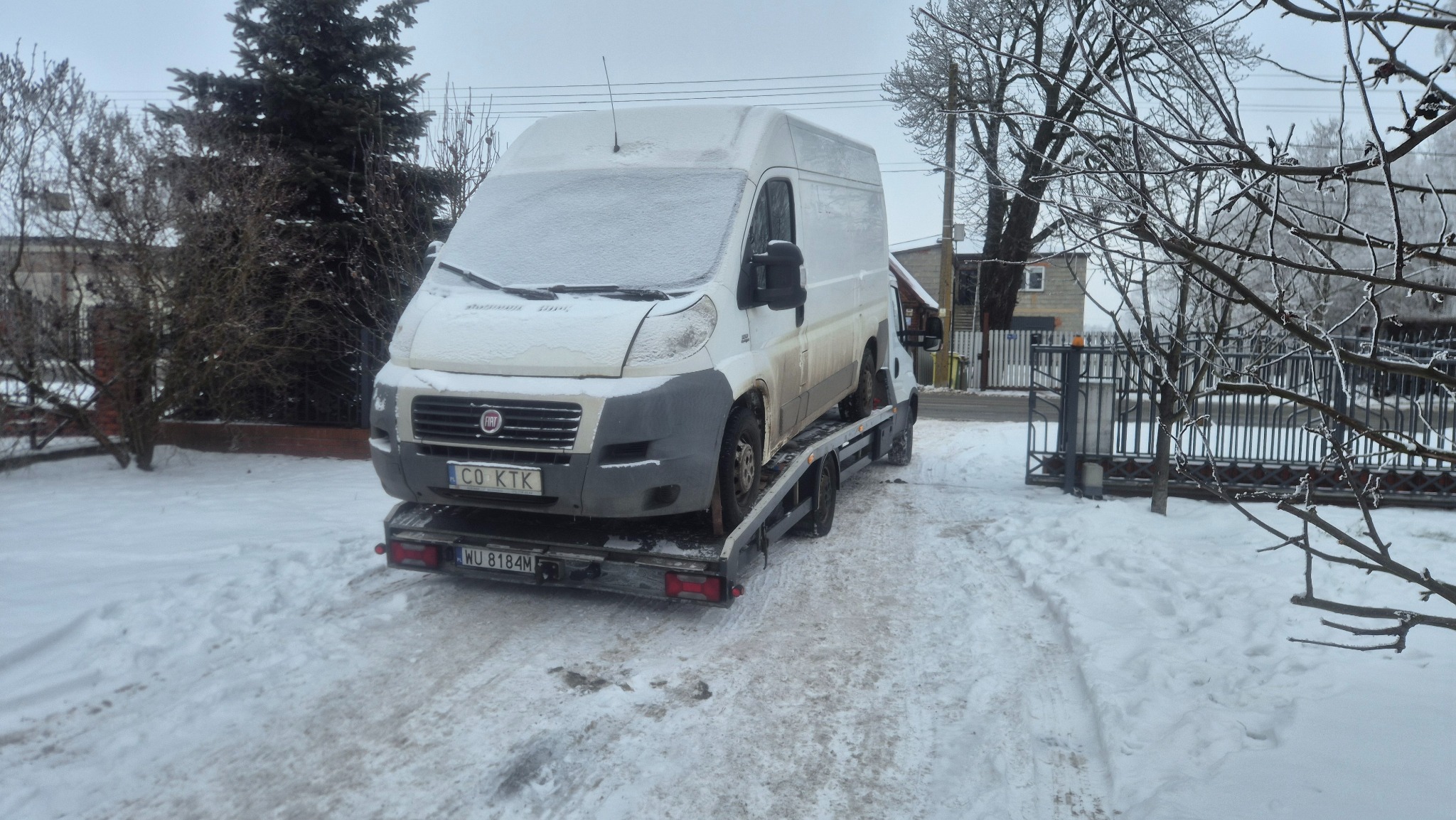 Biały bus Fiat na lawecie w zimowej scenerii. Śnieg pokrywa pojazd i otoczenie, widoczne drzewa i budynek w tle. Tablice rejestracyjne widoczne.