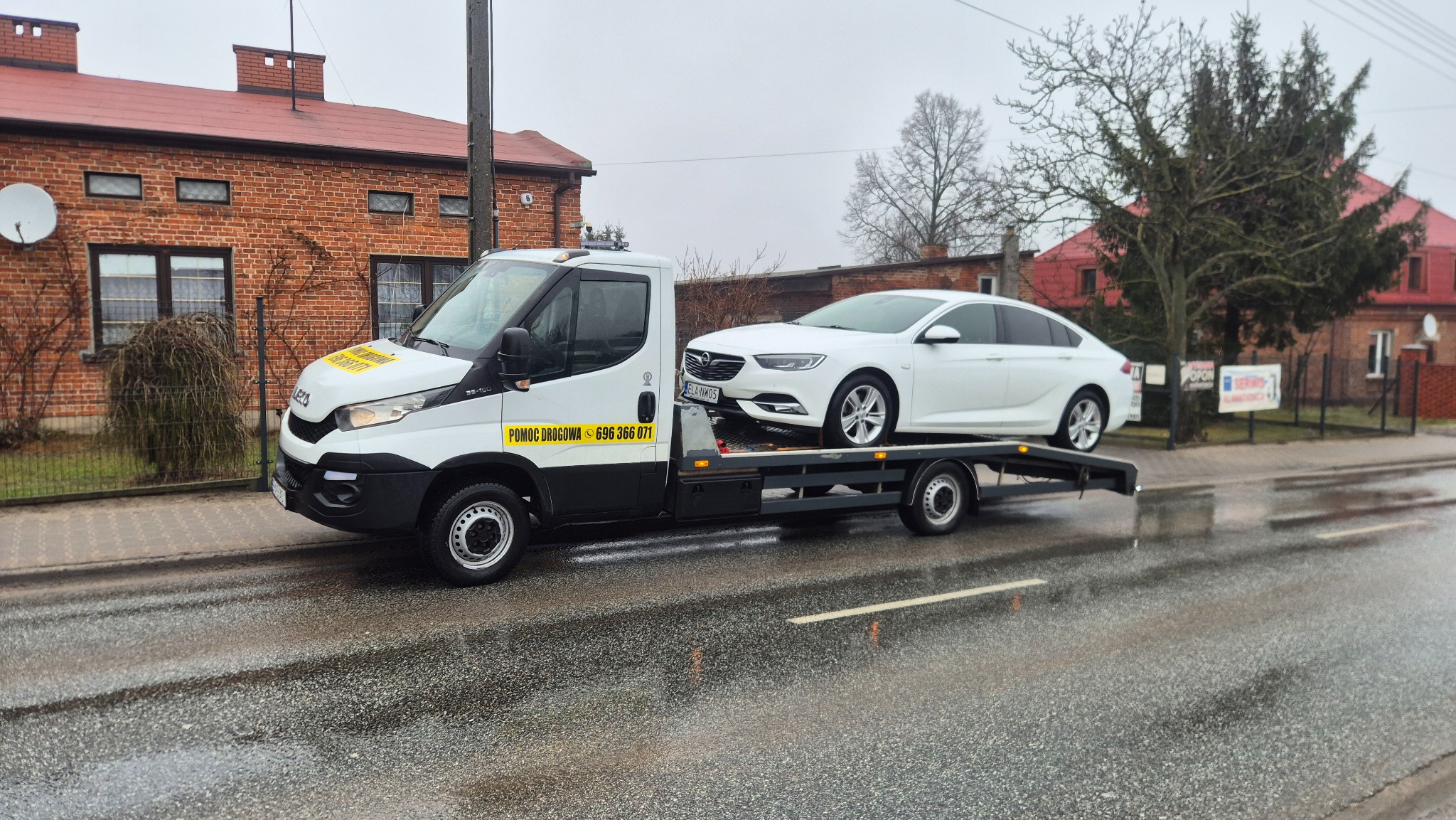 Biała laweta Iveco z białym samochodem osobowym Opel Insignia na platformie, na tle ceglanego budynku i mokrej nawierzchni drogi.