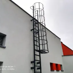 Metalowa drabina ewakuacyjna z koszem ochronnym, przymocowana do jasnoszarej ściany budynku z czerwonymi akcentami architektonicznymi.