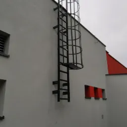 Zewnętrzna metalowa drabina ewakuacyjna z klatką bezpieczeństwa, przymocowana do szarej ściany budynku z czerwonymi akcentami architektonicznymi.