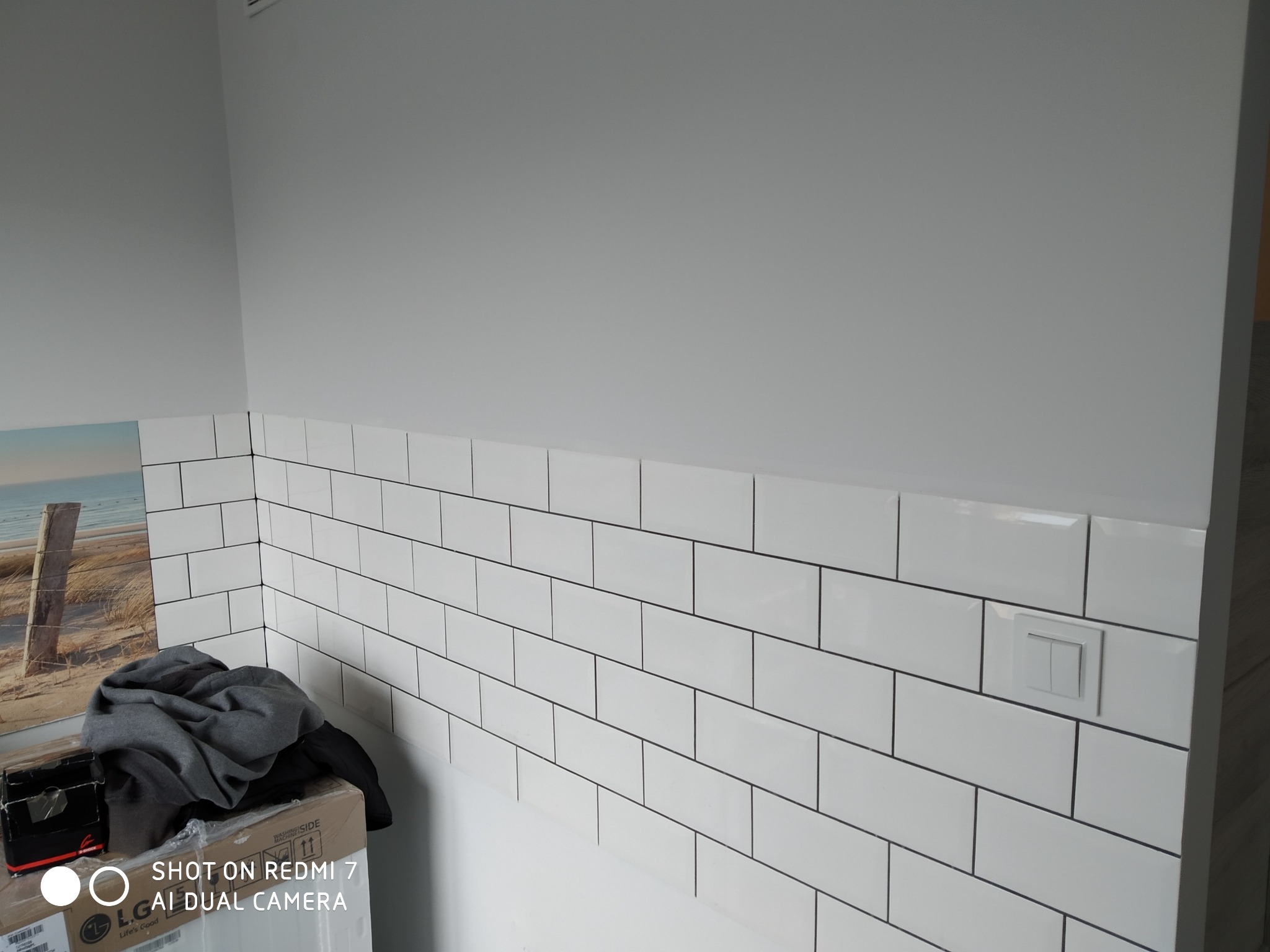 Ściana w łazience wyłożona białymi, prostokątnymi płytkami, z widocznym włącznikiem światła i fragmentem dekoracji ściennej z motywem plaży, na pierwszym planie karton z ubraniami.