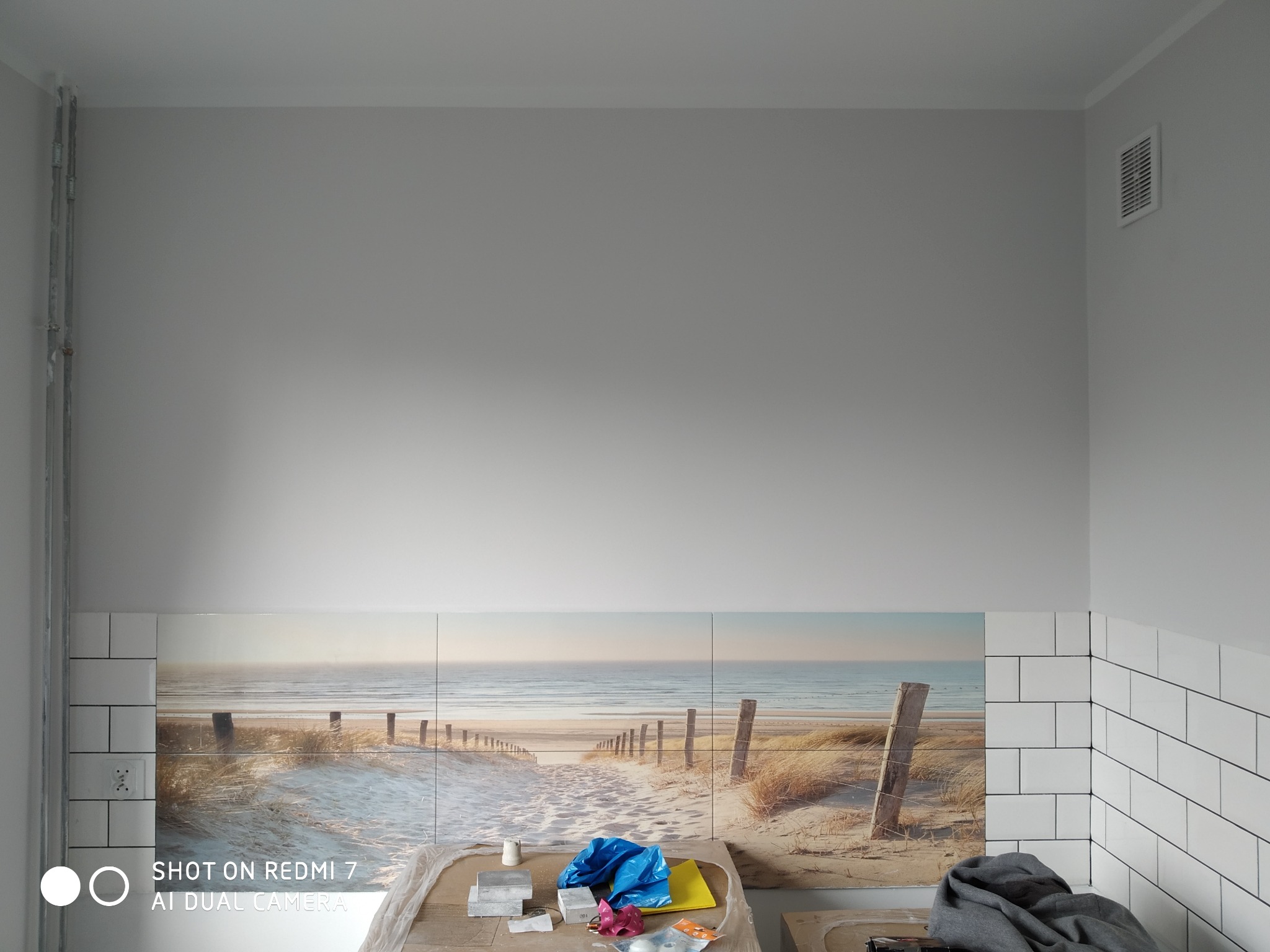 Ściana nad blatem w trakcie wykańczania z białymi kaflami i fototapetą z motywem plaży, widoczne narzędzia i materiały budowlane na blacie.