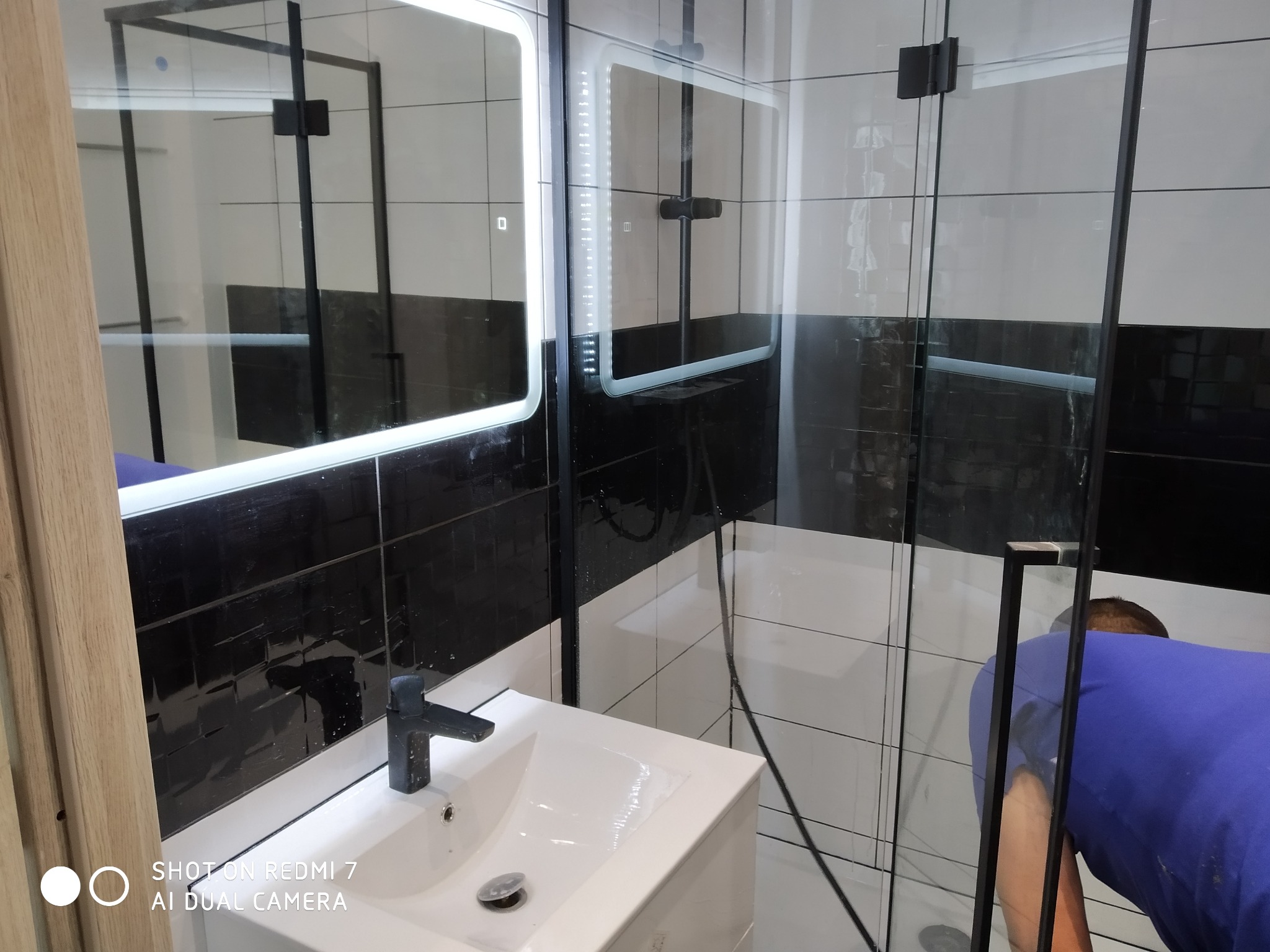 Nowoczesna łazienka z czarno-białymi płytkami, podświetlanym lustrem i szklaną kabiną prysznicową; osoba w niebieskiej koszulce czyści kabinę.