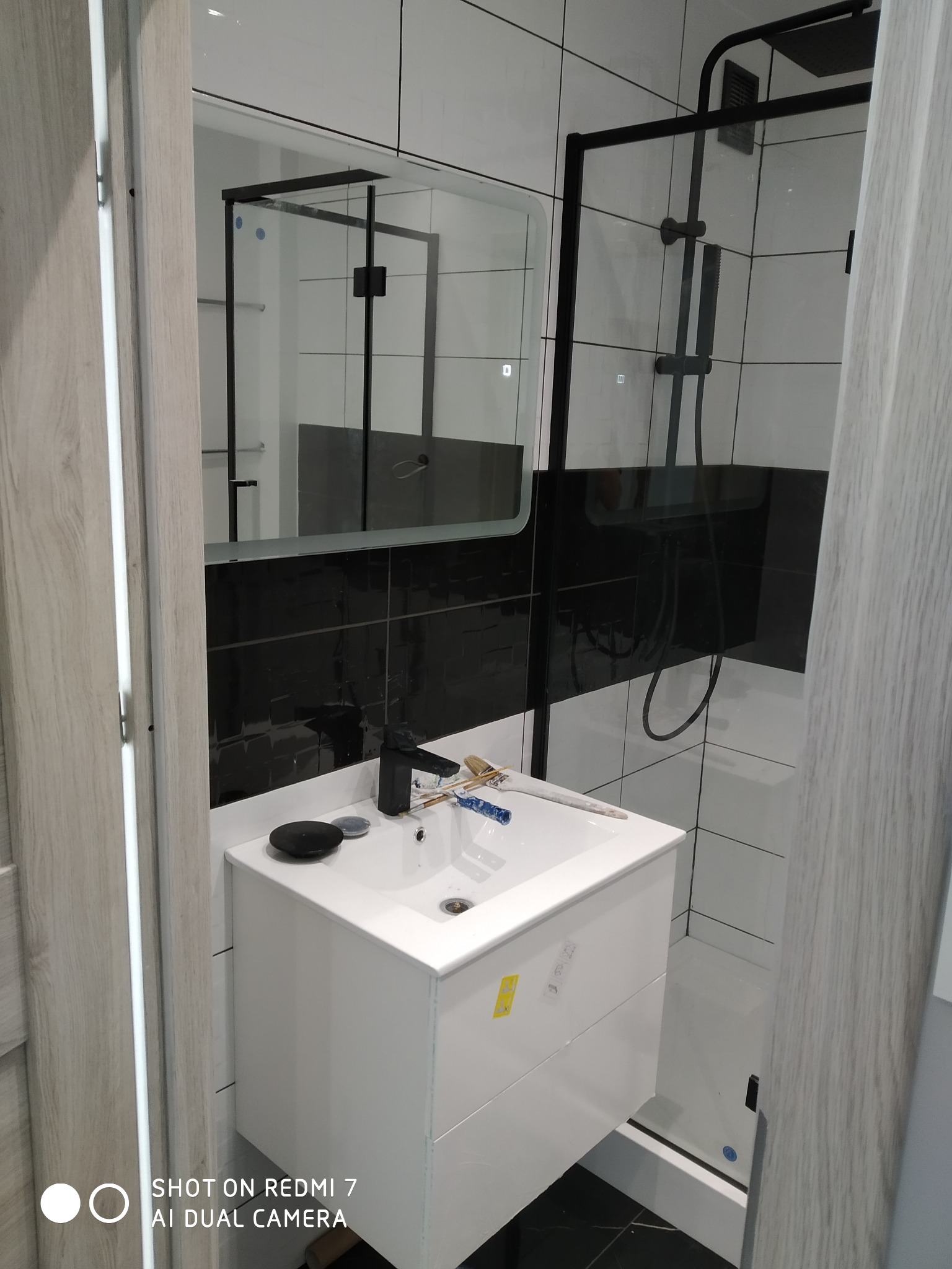 Wykończona łazienka z czarno-białymi płytkami, kabiną prysznicową typu walk-in z czarną armaturą i białą szafką pod umywalkę z czarną baterią, widoczne narzędzia malarskie na umywalce.