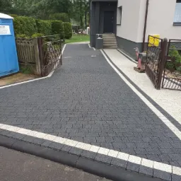 Nowo ułożona, grafitowa kostka brukowa z białymi pasami obramowania, prowadząca do domu z ciemnymi drzwiami i bramą, widoczna ubijarka wibracyjna i przenośna toaleta w tle.