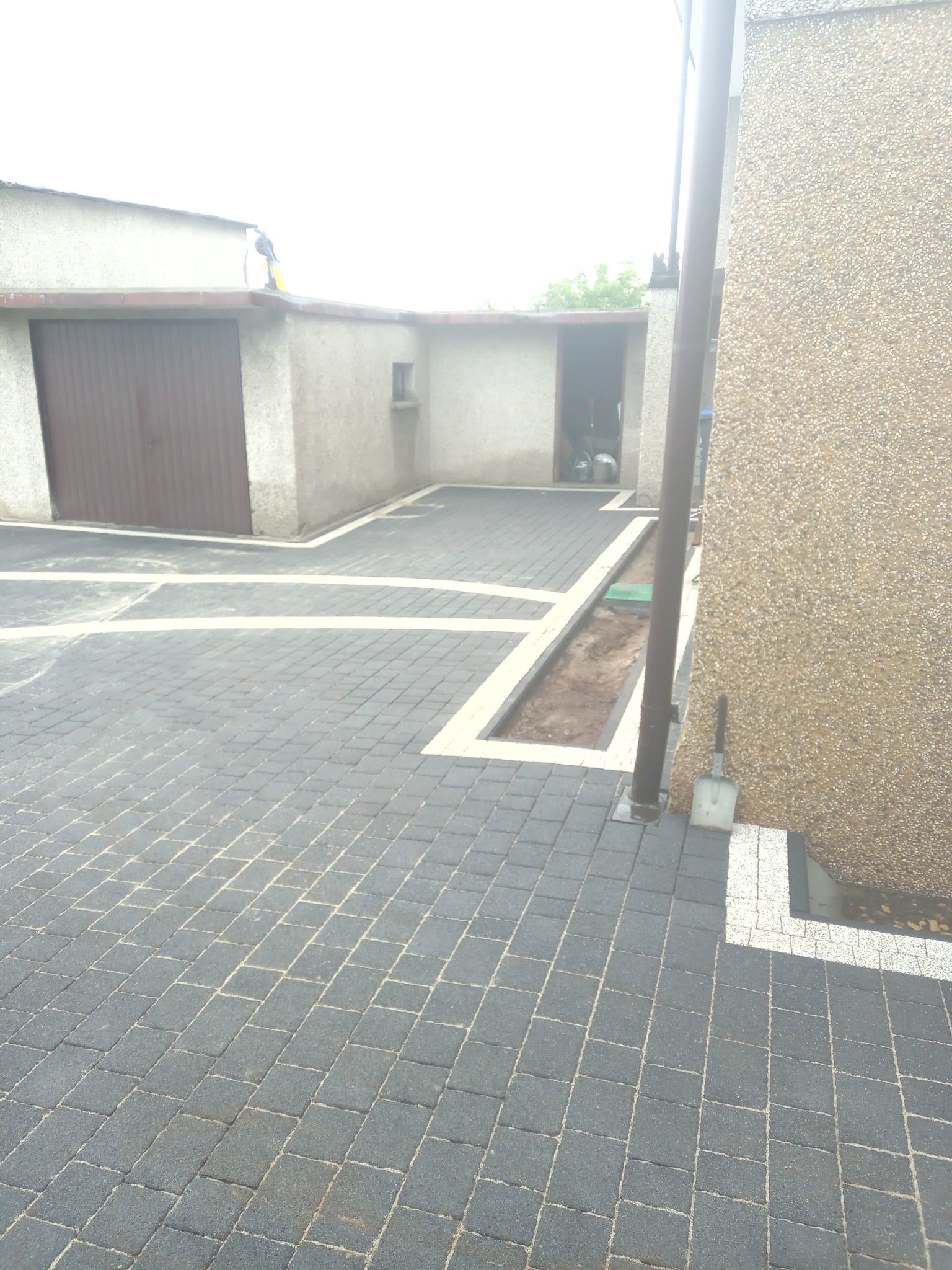 Ułożona szara kostka brukowa z białymi obrzeżami tworząca wzór krzyża na podjeździe, widoczna łopata oparta o ścianę budynku.