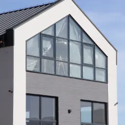 Nowoczesny dom z dużym trójkątnym oknem na poddaszu, zabezpieczonym folią ochronną, widoczna drabina wewnątrz, poniżej dwa mniejsze okna z szarymi ramami.