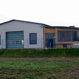 Nowoczesny budynek przemysłowy z szarą fasadą, niebieskimi ramami okiennymi i bramą segmentową, otoczony zielenią i ogrodzeniem z siatki.