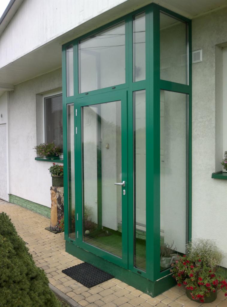 Szklana zabudowa wejścia do domu z zielonymi profilami, widoczny dywanik i donice z kwiatami, na ścianie dekoracja z drewna.