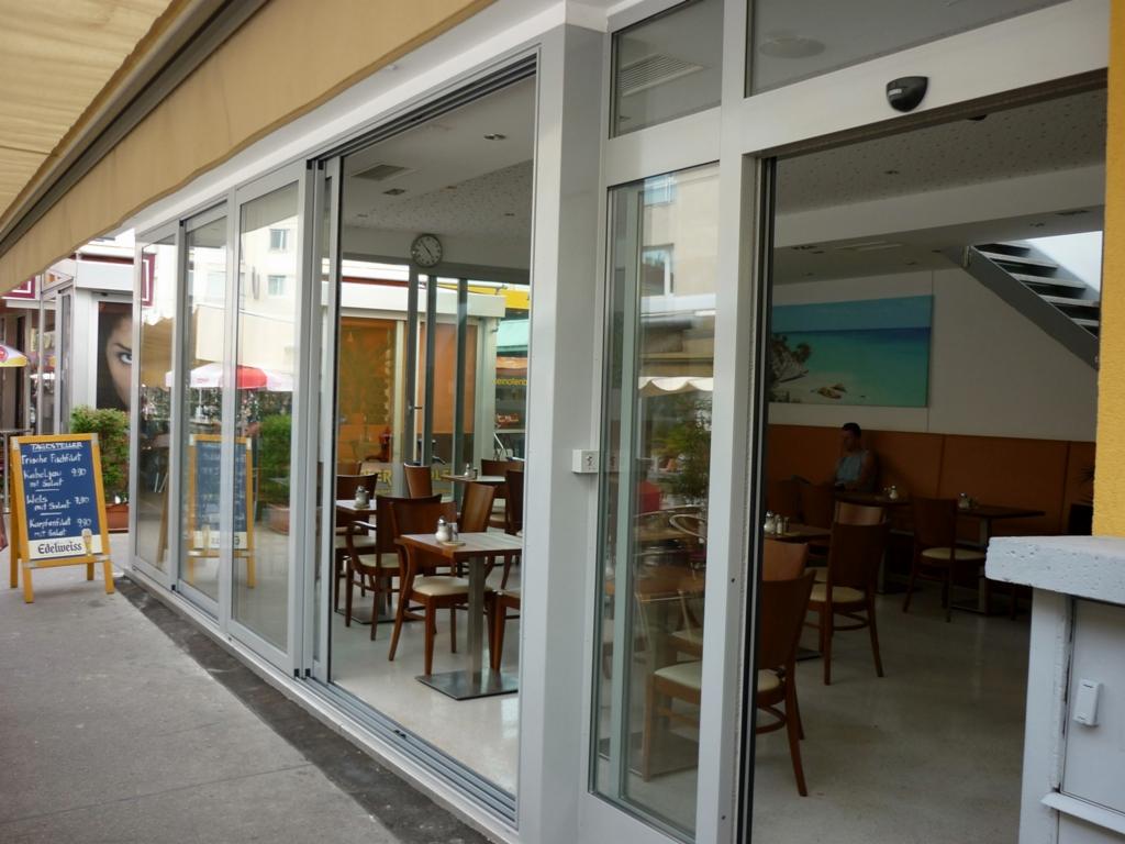 Szerokie, przeszklone drzwi przesuwne z aluminium, oddzielające wnętrze kawiarni od chodnika, widoczne menu na tablicy przed wejściem.