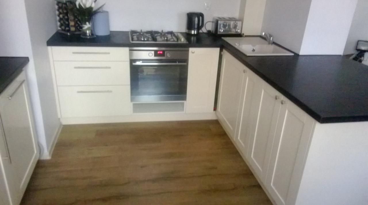 Jasna zabudowa kuchenna z ciemnym blatem, widoczna płyta grzewcza i piekarnik, drewniana podłoga.