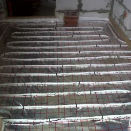 Białe rurki ogrzewania podłogowego, przymocowane czarnymi spinkami do folii z czerwoną kratką, ułożone w surowym pomieszczeniu z betonowymi ścianami.