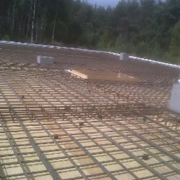 Zbrojenie płyty stropowej z widocznymi dystansami, betonowymi podkładkami i deskowaniem otworu, przygotowane do zalania betonem, w tle las.