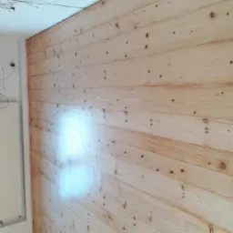 Ściana wyłożona jasnymi, surowymi panelami drewnianymi z widocznymi sękami, świeżo po montażu, z fragmentem okna i grzejnika w tle.