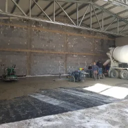 Wnętrze hali budowlanej podczas wylewania i wygładzania posadzki. Widoczny betonowóz, ekipa pracująca oraz zacieraczki do betonu.