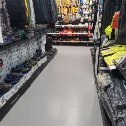 Wnętrze sklepu budowlanego: widok na alejkę z odzieżą roboczą i obuwiem ochronnym marki Hogert, narzędzia glazurnicze Hardy na p&oacute;łkach w tle, posadzka w kolorze jasnoszarym.