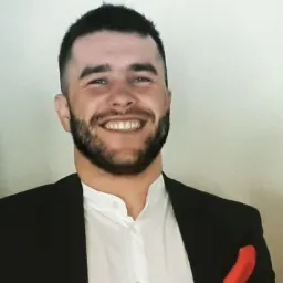 Uśmiechnięty mężczyzna z brodą w czarnej marynarce i białej koszuli, z czerwoną poszetką w kieszeni. Portret w jasnym otoczeniu, lekko z prawej strony kadru.